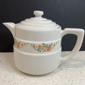 May include: White ceramic teapot with a lid and handle. The teapot features a floral design with pink, yellow, and green flowers. The teapot is sitting on a white surface.