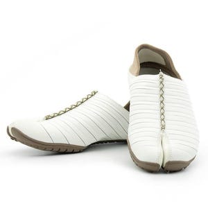 May include: A pair of white tabi shoes featuring a split-toe design, a series of horizontal stitched panels across the upper, and a central olive green lace-up detail. The shoes have a tan interior lining and a textured brown rubber sole.