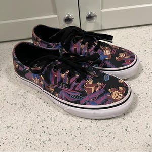 May include: Pair of Vans trainers featuring a black, purple, and tan floral design. The shoes have black laces, a white sole, and the Vans logo. The trainers are on a light-coloured surface.
