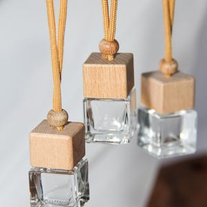 May include: Three hanging car air fresheners. Each has a clear, square glass bottle topped with a light brown wooden block and a wooden bead. The bottles hang from a tan cord.