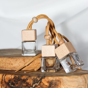 Peut inclure: Trois désodorisants pour voiture en verre transparent avec des dessus en bois et des cordons beiges sont présentés sur une planche à découper en bois. Les désodorisants ont une forme carrée et sont partiellement remplis de liquide.