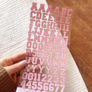 May include: A sheet of pink and gold alphabet and number stickers. The stickers are in a bold, block letter font with a gold outline. The letters and numbers are in various sizes and are arranged in rows. The background is a light brown wood surface.