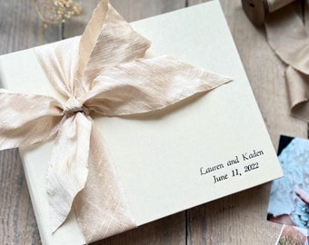 May include: A white linen wedding album with a large beige silk ribbon tied in a bow. The album is personalized with the text "Lauren and Kaden June 11, 2022".