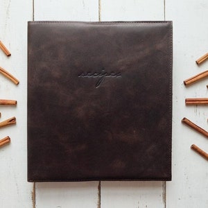 May include: A brown leather recipe binder with the word "recipes" embossed on the front. The binder is surrounded by cinnamon sticks on a white wooden background.