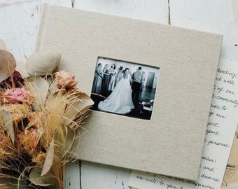 Puede incluir: Un álbum de fotos cubierto de lino con una ventana cuadrada que muestra una foto en blanco y negro de una ceremonia de boda. Un ramo de flores secas con rosas rosas y hojas de eucalipto está en primer plano.