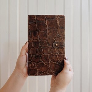 May include: A brown leather-bound journal with a textured, alligator-like pattern. The journal has a brass button closure.