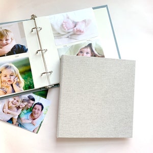 Puede incluir: Un álbum de fotos cuadrado con una cubierta de lino gris claro. El álbum abierto cuenta con un mecanismo de encuadernación de tres anillas metálicas que sujeta varias fotografías, incluyendo imágenes de niños, pies de bebé y un retrato familiar.