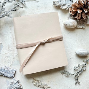 May include: A light beige leather journal with a blush pink velvet ribbon tied around it. The journal is on a white background with decorative elements.