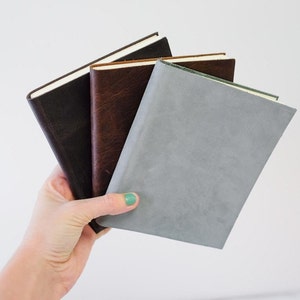 May include: Three leather-bound journals in different colors: dark brown, light brown, and gray. The journals are stacked on top of each other.