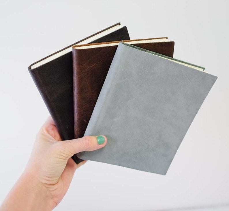 May include: Three leather-bound journals in different colors: dark brown, light brown, and gray. The journals are stacked on top of each other.
