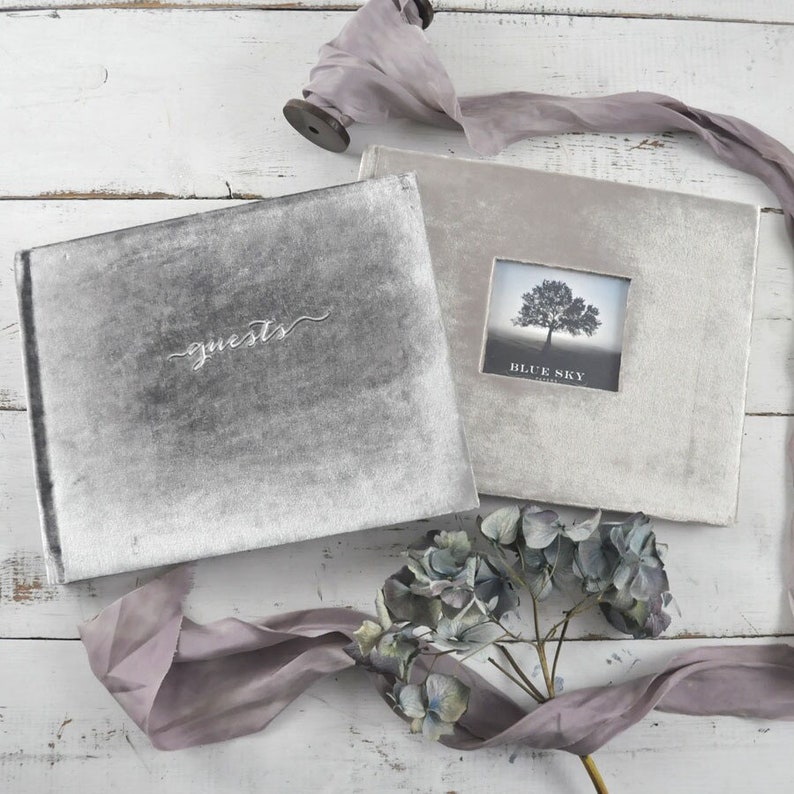 May include: Two gray velvet guest books with the words "guests" and "Blue Sky" printed on the covers. The books are on a white wooden surface with a purple ribbon and a bouquet of blue flowers.