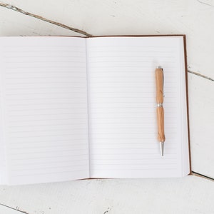 May include: An open lined notebook with a wooden pen on the right page. The notebook is on a white wooden surface.