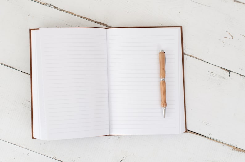 May include: An open lined notebook with a wooden pen on the right page. The notebook is on a white wooden surface.