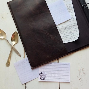 May include: A brown leather recipe binder with a white recipe card on a white wooden surface. The recipe card has a handwritten recipe with the words "Serves" and "Ingredients".
