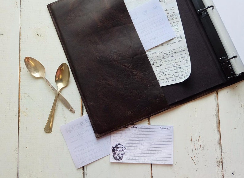 May include: A brown leather recipe binder with a white recipe card on a white wooden surface. The recipe card has a handwritten recipe with the words "Serves" and "Ingredients".