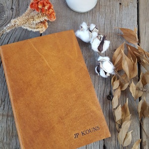 May include: A brown leather-bound journal with the name "JP KOUNS" embossed on the front cover. The journal is sitting on a wooden surface with dried flowers and a candle.
