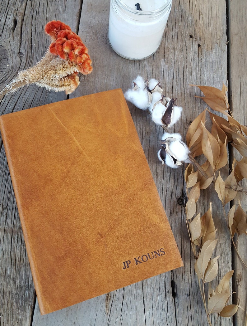 May include: A brown leather-bound journal with the name "JP KOUNS" embossed on the front cover. The journal is sitting on a wooden surface with dried flowers and a candle.