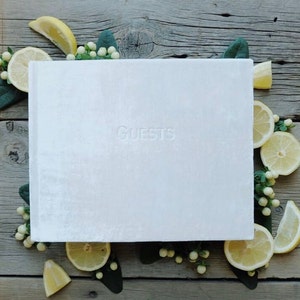 May include: White velvet guest book with the word "GUESTS" embossed on the front cover. The book is surrounded by lemons and white flowers.