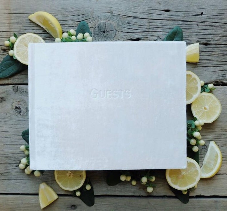 May include: White velvet guest book with the word "GUESTS" embossed on the front cover. The book is surrounded by lemons and white flowers.