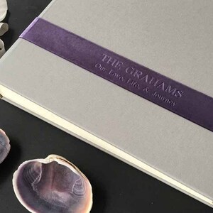 Puede incluir: Un libro de tapa dura gris con una banda de terciopelo morado oscuro sobre la cubierta. La banda tiene grabado el texto THE GRAHAMS Our Love, Life, & Journey. El libro est&aacute; colocado junto a dos conchas marinas abiertas y una cinta blanca.