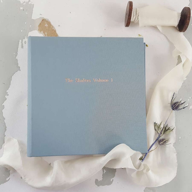 Puede incluir: Un diario de tapa dura de color azul claro con el t&iacute;tulo "The Stories Volume 1" en relieve en letras doradas. El diario est&aacute; sobre una superficie blanca con una cinta blanca y flores secas.