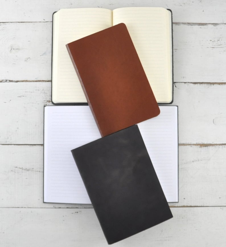 May include: Three notebooks with lined pages. The top notebook is cream colored with brown leather cover. The middle notebook is brown leather with a cream colored cover. The bottom notebook is black leather with a cream colored cover.