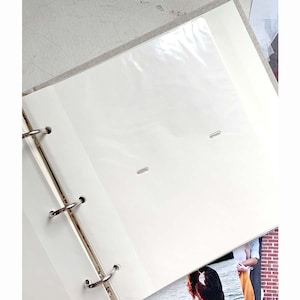 May include: Open photo album with clear plastic sleeves and white pages. The album is bound with silver metal rings and has a light beige fabric cover. A photograph is visible in the bottom right corner.