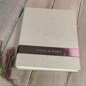 Puede incluir: Un &aacute;lbum de fotos rectangular con una cubierta de lino beige claro. Una cinta horizontal de color granate oscuro envuelve el centro, con el texto NANA & PAPA grabado en un acabado met&aacute;lico. El &aacute;lbum tiene un dise&ntilde;o limpio y estructurado.