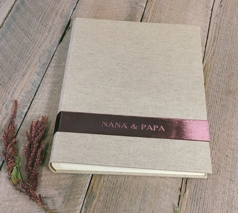 Puede incluir: Un &aacute;lbum de fotos rectangular con una cubierta de lino beige claro. Una cinta horizontal de color granate oscuro envuelve el centro, con el texto NANA & PAPA grabado en un acabado met&aacute;lico. El &aacute;lbum tiene un dise&ntilde;o limpio y estructurado.
