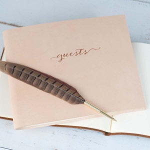May include: A light pink leather guest book with the word "guests" written in cursive on the front. A brown and black feather pen is resting on the book.