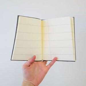 May include: A gray, lined notebook with a soft cover is open to a blank page. The notebook is being held by a hand.