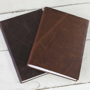 May include: Two leather-bound journals, one brown and one dark brown, are stacked on top of each other. The journals have a simple, elegant design and are perfect for writing, sketching, or journaling.