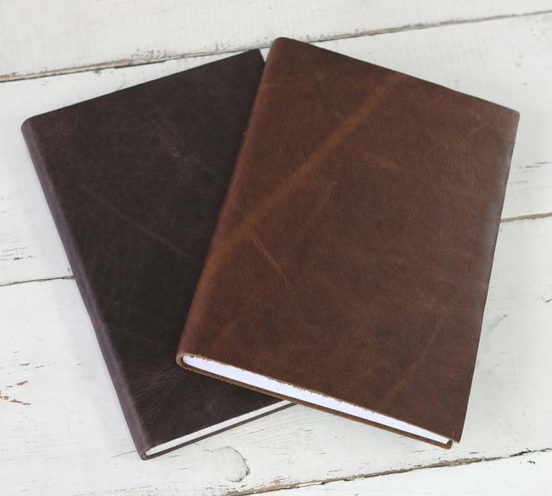 May include: Two leather-bound journals, one brown and one dark brown, are stacked on top of each other. The journals have a simple, elegant design and are perfect for writing, sketching, or journaling.