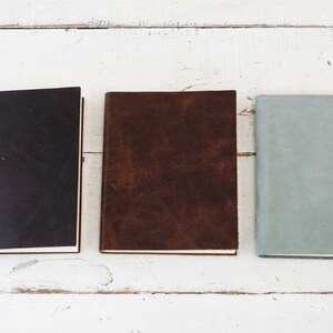 May include: Three closed notebooks with different colored covers. The notebook on the left has a black cover, the middle notebook has a brown cover, and the notebook on the right has a light blue cover.