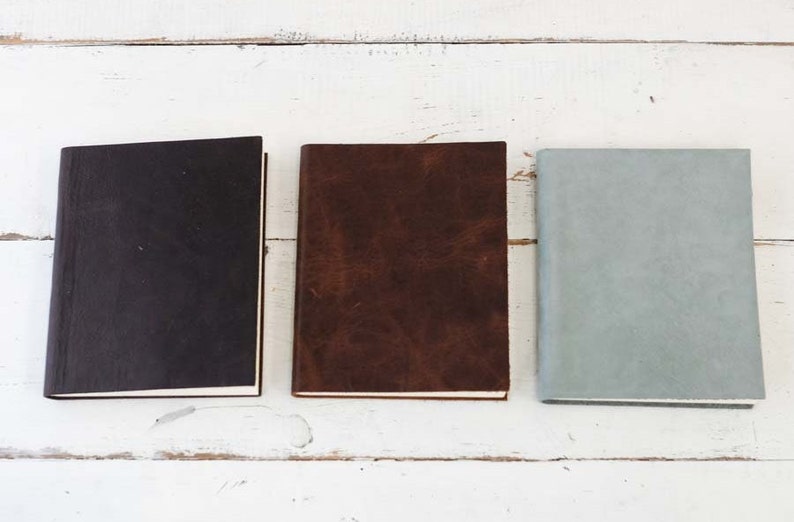 May include: Three closed notebooks with different colored covers. The notebook on the left has a black cover, the middle notebook has a brown cover, and the notebook on the right has a light blue cover.