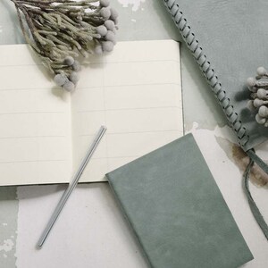 May include: Two green notebooks with a silver pen on a white surface. One notebook has a braided edge and the other has a plain cover.