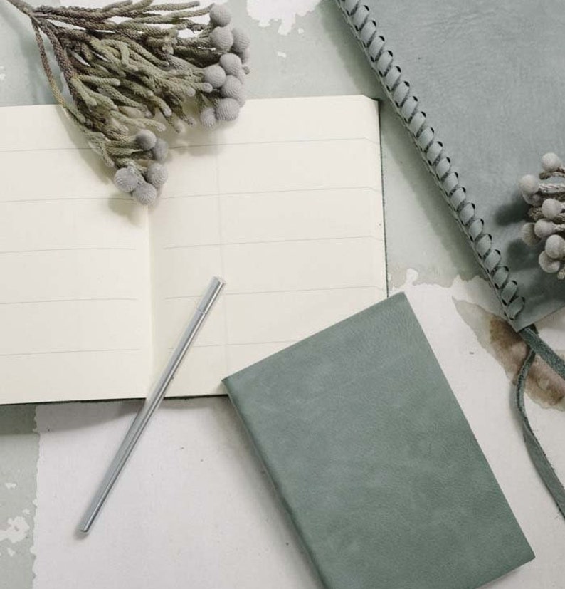 May include: Two green notebooks with a silver pen on a white surface. One notebook has a braided edge and the other has a plain cover.