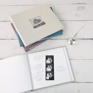May include: A white guest book with a clear plastic cover, open to a page with handwritten messages and a strip of black and white photo booth photos. The book is on a white wooden surface. The book cover has a small window with a photo of a tree and the text "Blue Sky" printed on it.