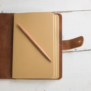 May include: A brown leather-bound journal with a gold pen lying on the open pages. The journal has a strap closure and is open to reveal blank pages.