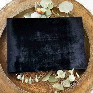 May include: A black velvet clutch with a gold zipper. The clutch is sitting on a wooden tray with green leaves.