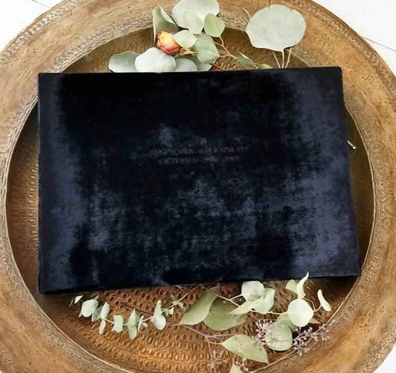 May include: A black velvet clutch with a gold zipper. The clutch is sitting on a wooden tray with green leaves.