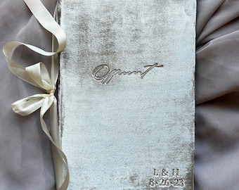 May include: A silver velvet guest book with the word "Guest" embossed on the front. The book is tied with a white satin ribbon and has the initials "L & H" and the date "8.26.23" embossed on the bottom right corner.