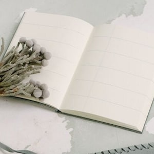 May include: A blank, lined notebook with a gray cover is open on a white surface. A sprig of dried, gray flowers is placed on the open pages.