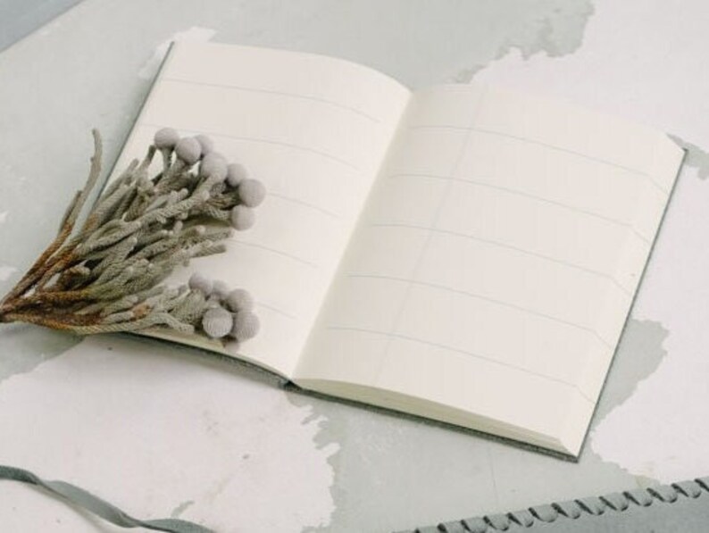 May include: A blank, lined notebook with a gray cover is open on a white surface. A sprig of dried, gray flowers is placed on the open pages.