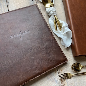 May include: A brown leather recipe binder with the word "recipes" embossed on the front. The binder is closed and lying on a light wood surface.