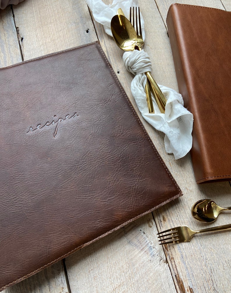 May include: A brown leather recipe binder with the word "recipes" embossed on the front. The binder is closed and lying on a light wood surface.