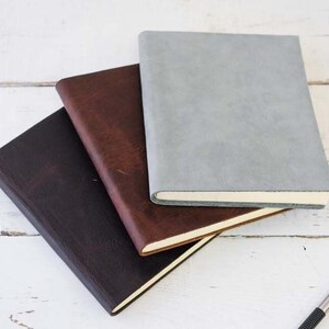 May include: Three leather-bound journals stacked on top of each other. The top journal is light gray, the middle journal is brown, and the bottom journal is dark brown.