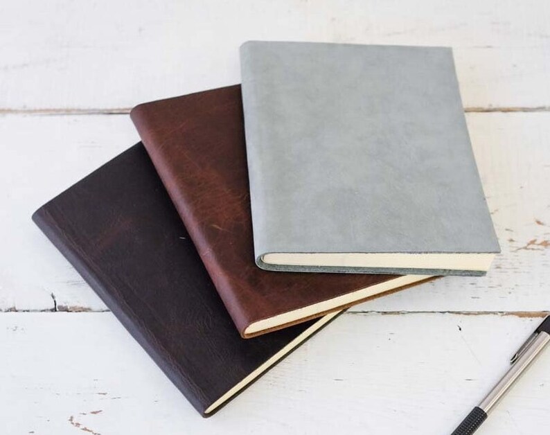 May include: Three leather-bound journals stacked on top of each other. The top journal is light gray, the middle journal is brown, and the bottom journal is dark brown.