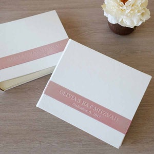 Puede incluir: Dos &aacute;lbumes de fotos rectangulares blancos con una banda horizontal de color rosa polvoriento en el centro. La banda est&aacute; grabada con el texto OLIVIA'S BAT MITZVAH y la fecha 9 de septiembre de 2017 en una fuente de letras may&uacute;sculas.