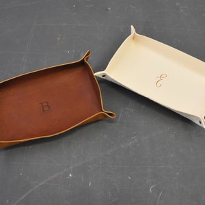 Puede incluir: Dos bandejas de cuero, una marrón y otra blanca, con las iniciales personalizadas "B" y "E" grabadas en la superficie.